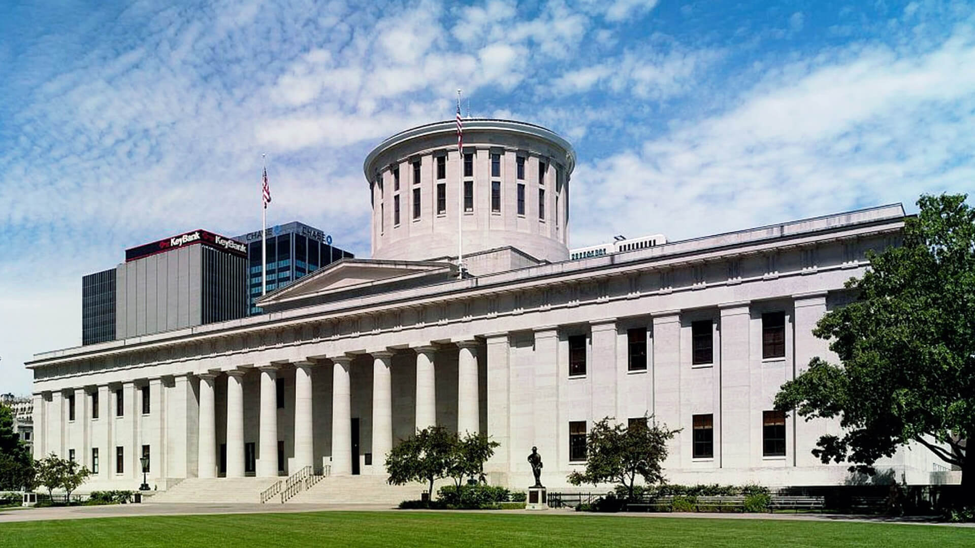 The Ohio Statehouse, a large, white, neoclassical building with a domed roof and a row of columns. The building is surrounded by a lawn and trees.