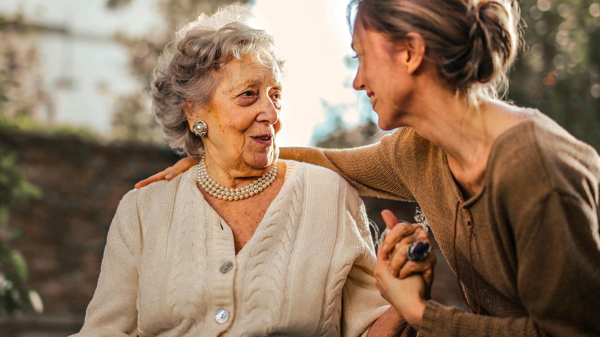 Two women sharing a warm moment outdoors; an elderly woman with pearl jewelry and white sweater smiles at a younger woman in beige who has her arm around her.