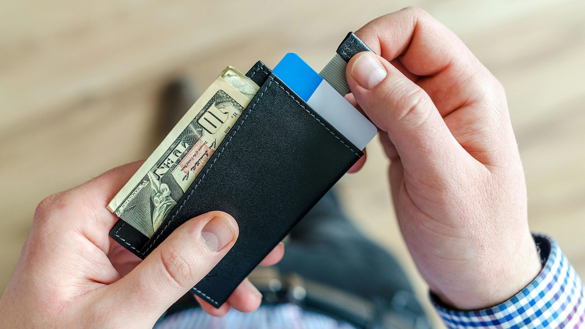 Hands holding a slim black leather wallet containing cash and a blue credit card, with the person wearing a blue checkered shirt sleeve visible.