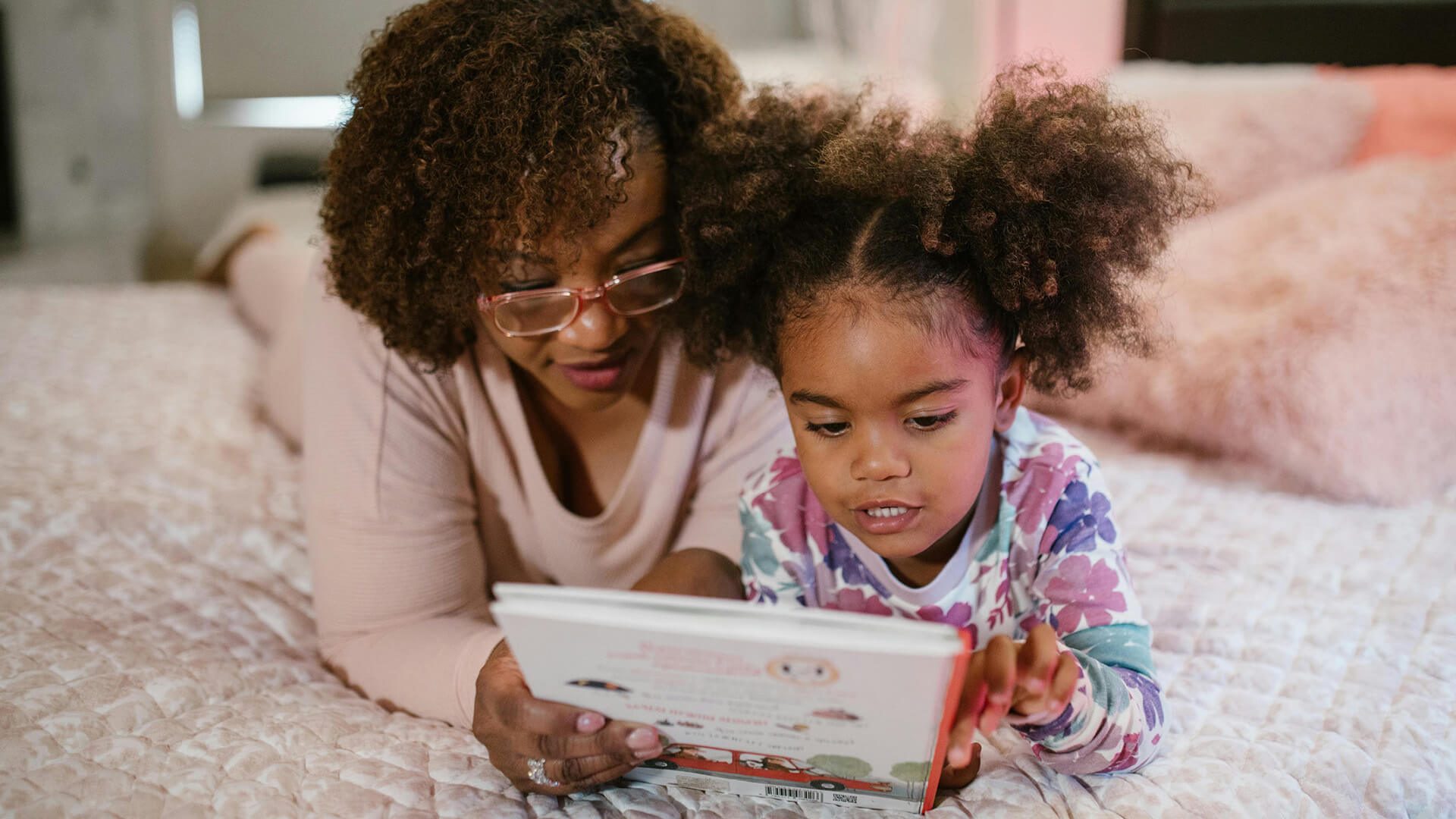 A mother and young daughter lying on a bed together, reading a children's book. Both have natural curly hair, and the mother wears glasses and a light pink sweater while the child wears a floral print top.